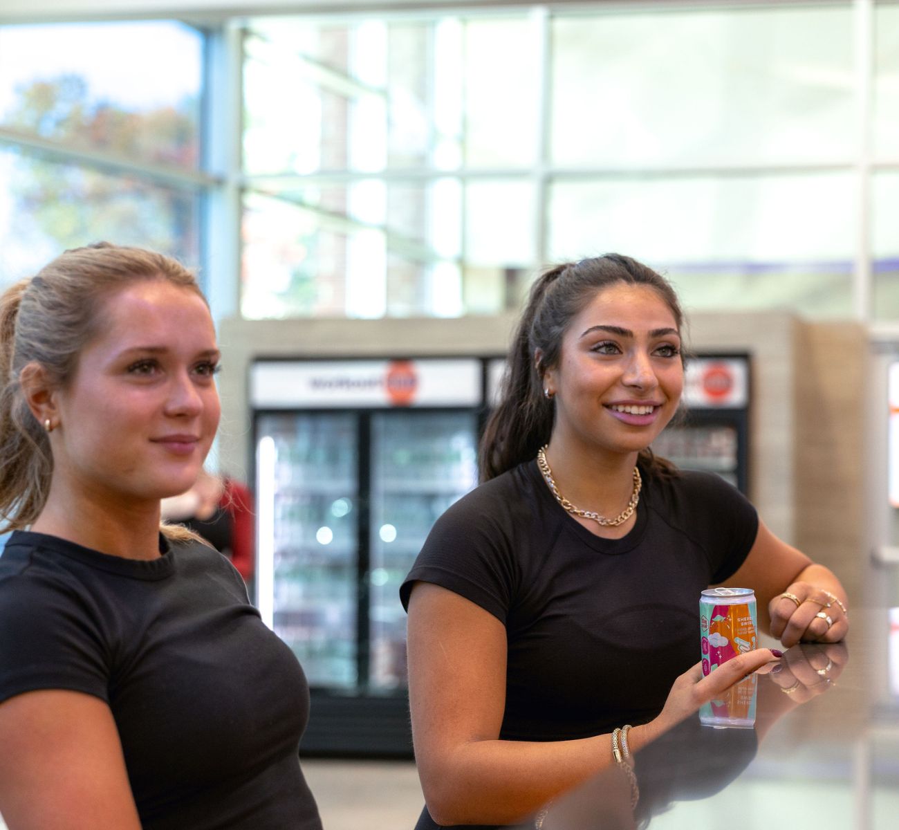 Two members relaxing and chatting at the shake bar counter at The Workout Club Salem NH with drinks and coolers in the background