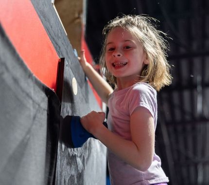Smiling young girl climbing an indoor rock wall at The Workout Club’s exciting and active kids day camp.