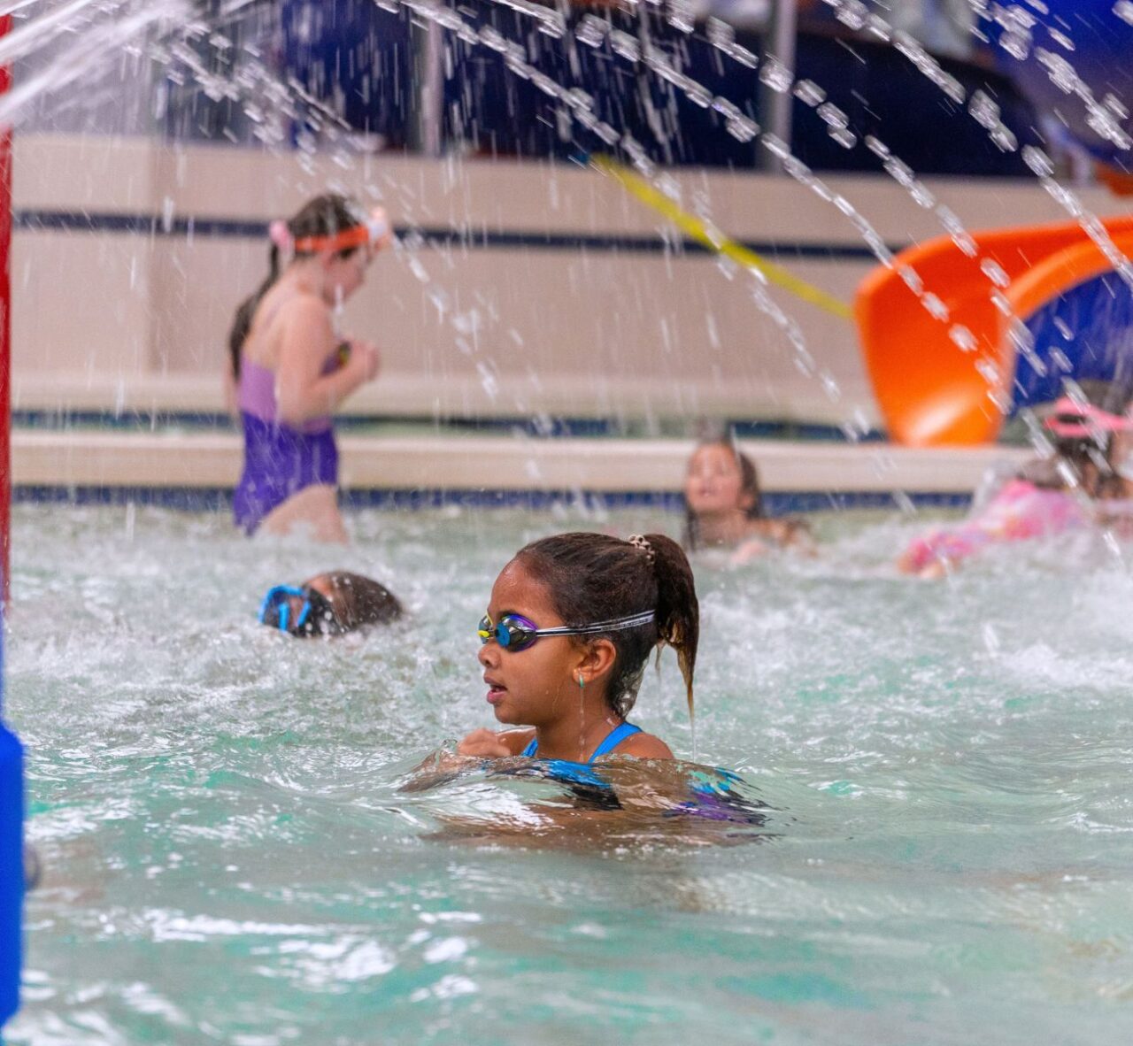 Girl swimming under water sprays in the indoor splash park pool during Kids Camp at The Workout Club Salem NH